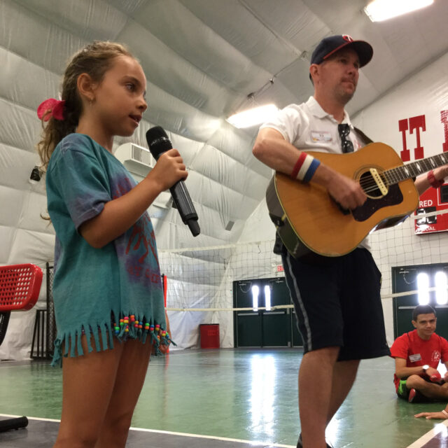 Girl camper singing into a microphone with a counselor playing the guitar at lineup