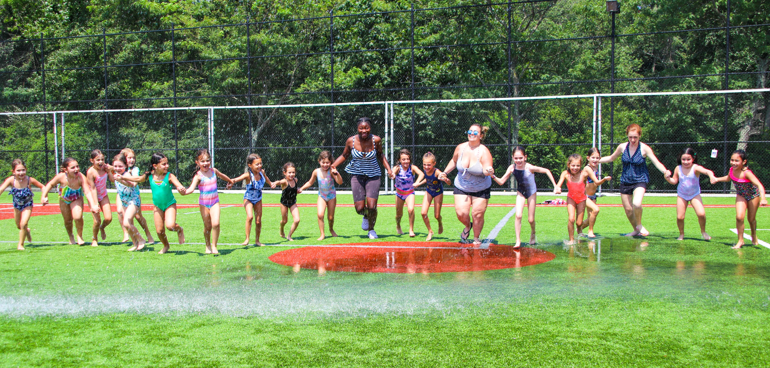 Campers and counselors hand in hand running through sprinklers.